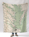 A person holding a blanket that features a vintage map design of the Virginia side of the Chesapeake Bay area with various town names marked in green on a beige background.