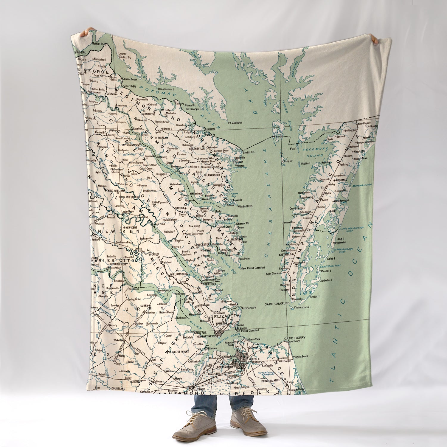 A person holding a blanket that features a vintage map design of the Virginia side of the Chesapeake Bay area with various town names marked in green on a beige background.